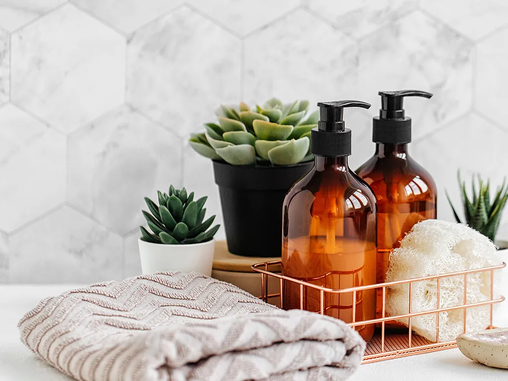 Butelki szamponu w koszyczku, obok ręcznik bawełniany i sztuczne kwiatki