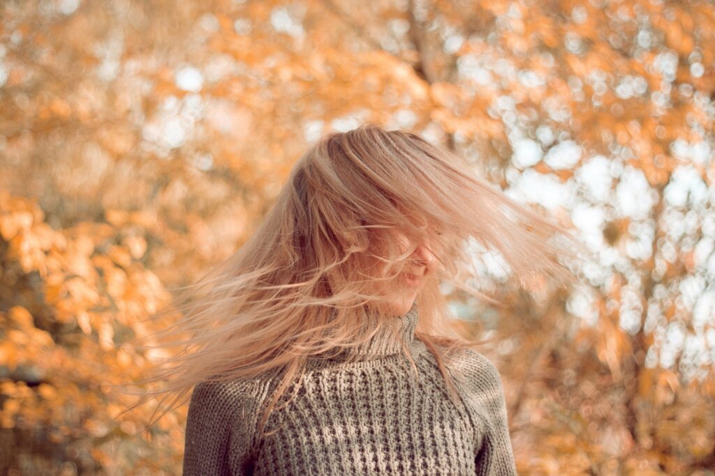 Kobieta kręci głową wprawiając w. ruch swoje blond włosy, w tle widać jesienne liście