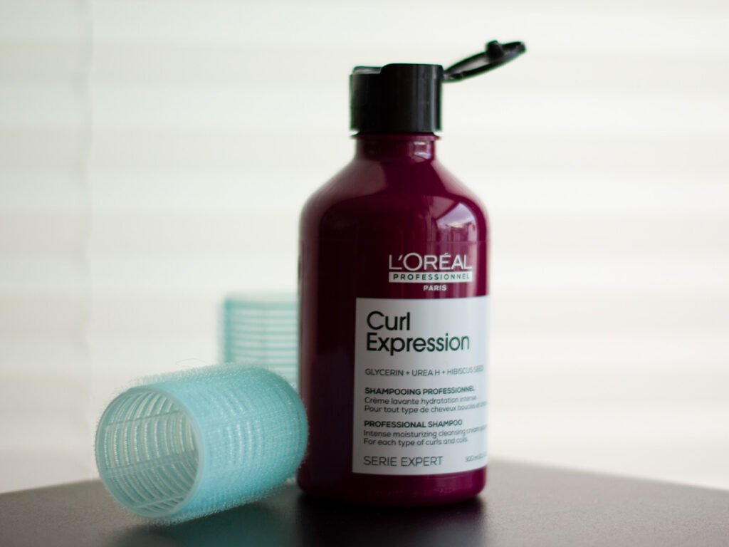 Szampon L’oreal Professionnel Curl Expression stojący na czarnej podstawce z wałkami do włosów - tytułowa.