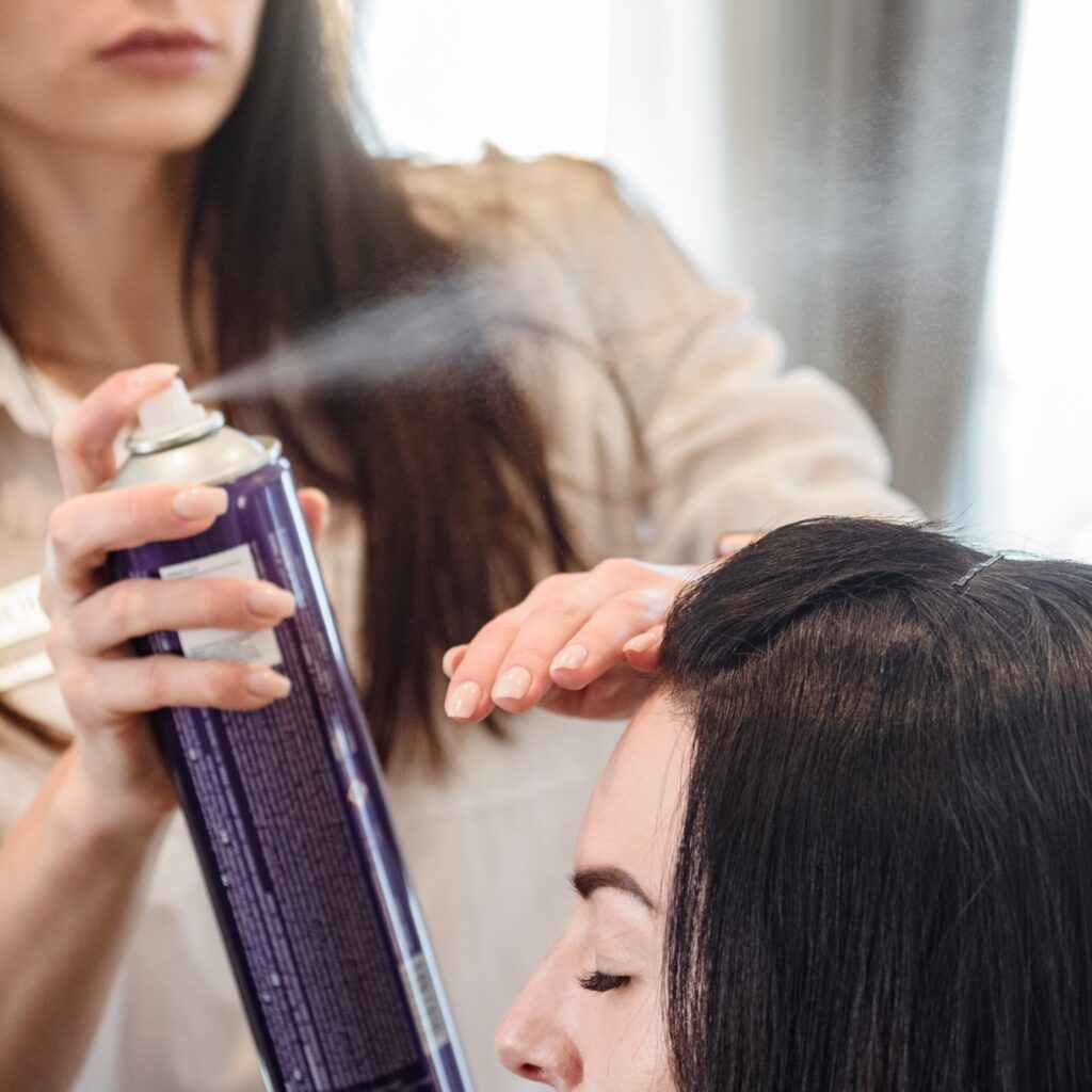 Fryzjerka aplikuje lakier do włosów na fryzurę pacjentki i chroni dłonią twarz przez lakierem