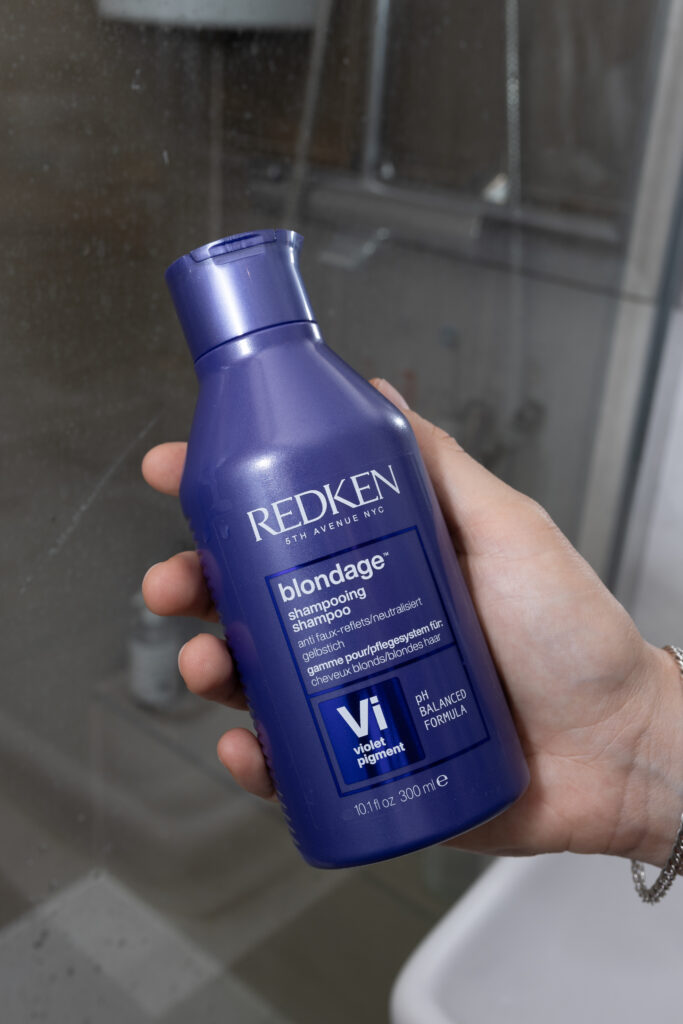 Kobieca dłoń trzymająca opakowanie szamponu Redken Color Extend Blondage, w tle prysznic