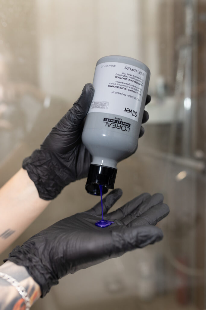 Kobiece dłonie w rękawiczce aplikują fioletowy szampon do siwych włosów L'Oréal Professionnel Silver na dłoń