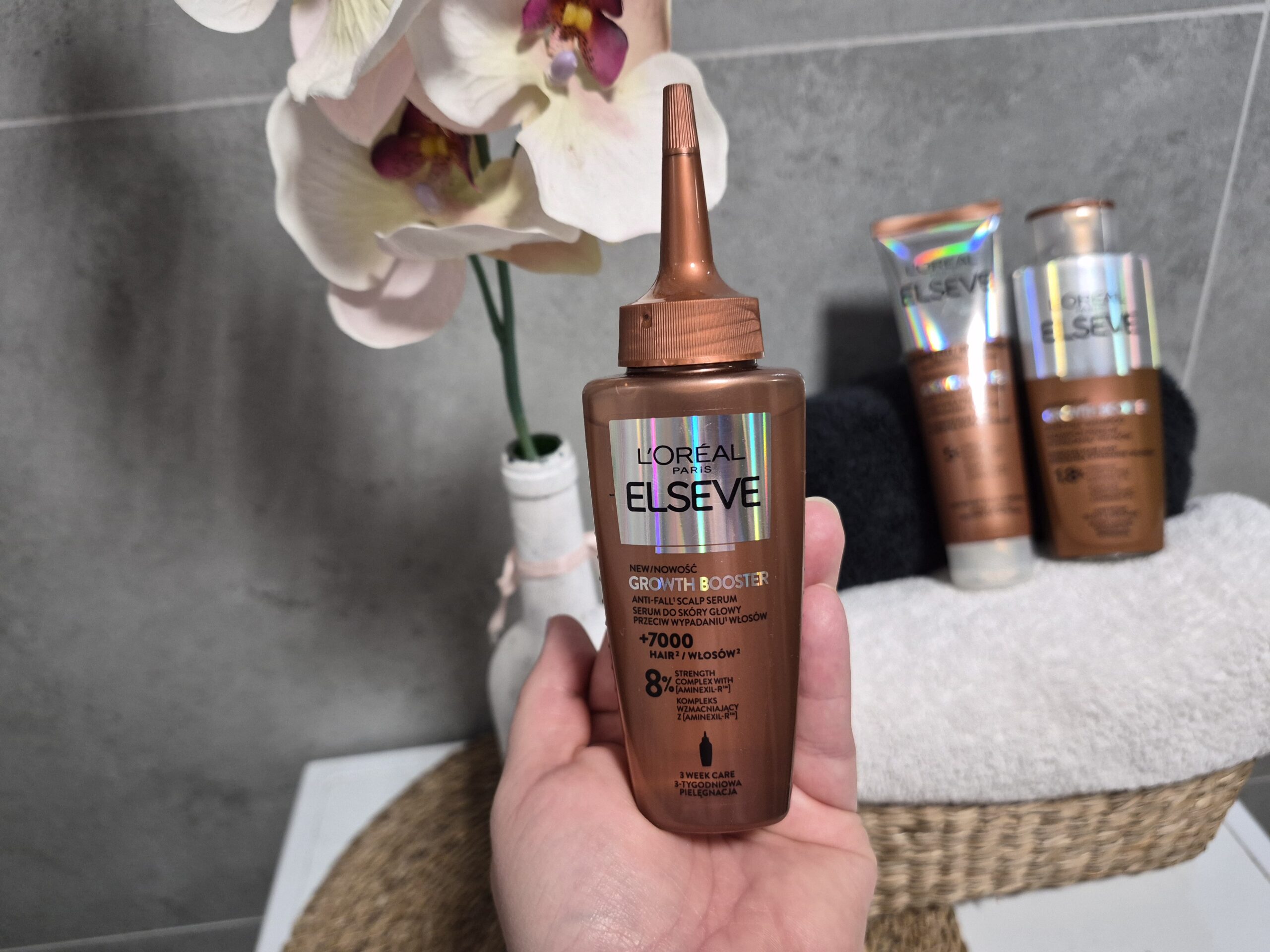 Kobieca dłoń w łazience trzyma opakowanie serum do skóry głowy przeciw wypadaniu włosów L’Oreal Paris Elseve Growth Booster