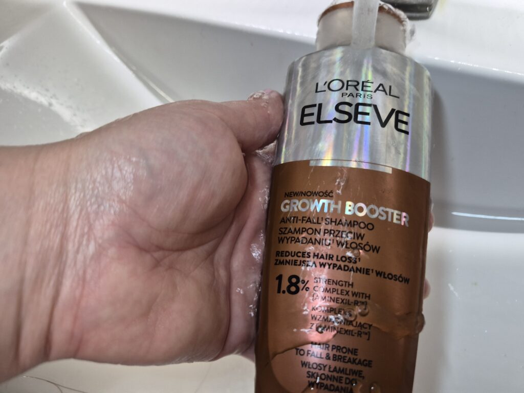 Kobieca dłoń trzyma pod bieżącą wodą szampon L’Oreal Paris Elseve Growth Booster