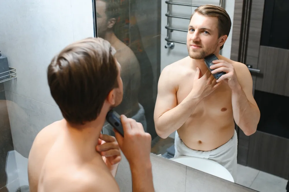 Mężczyzna przycina brodę trymerem przed łazienkowym lusterkiem 