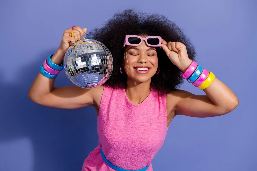 Młoda dziewczyna w różówym stroju, z kulą disco i włosami afro w stylu lat 70.